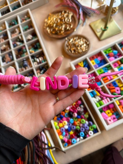 Pink chunky letter bead keychain spelling LINDIE in chunky alphabet beads, held over trays of letter beads