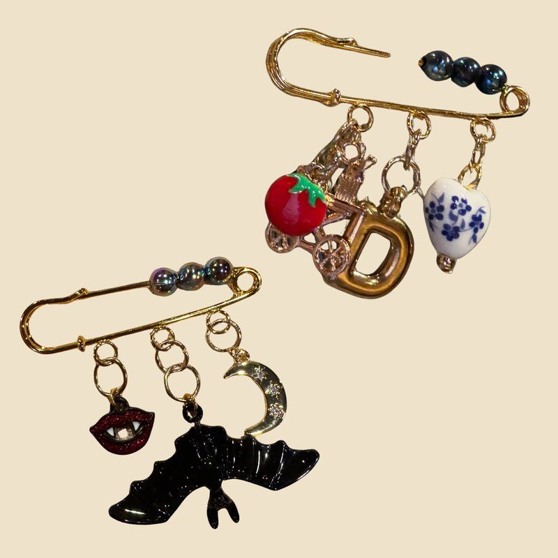 Two safety pin brooches with dangling charms, including a bat, a crescent moon, a tomato, a letter D and a porcelain heart