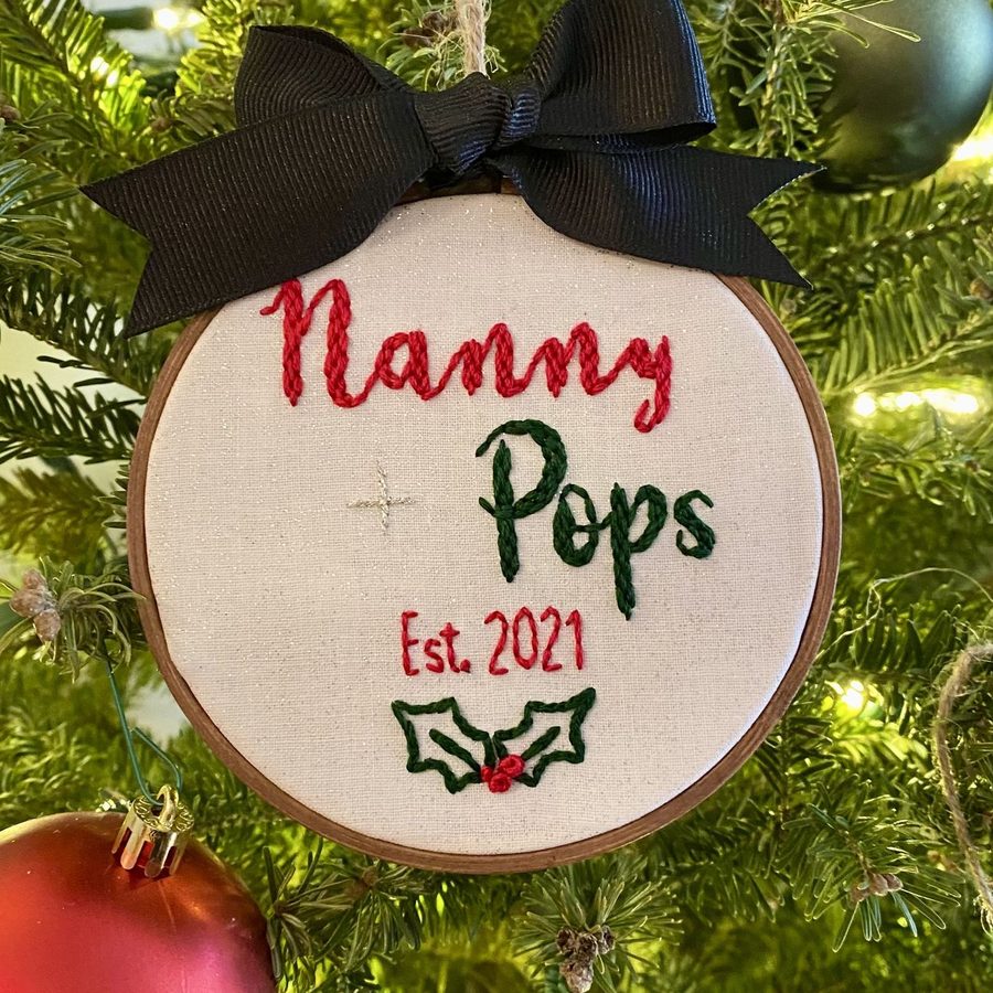 Hand-embroidered ornament reading Nanny + Pops, Est. 2021, with holly, hanging on a Christmas tree